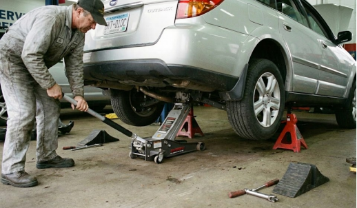 Mechanic lifting a silver car with a floor jack and using red jack stands for support.
