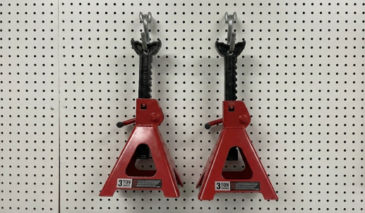 Two red 3-ton jack stands hanging neatly on a white garage pegboard.
